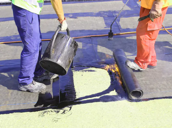 Two workers pouring hot bitumen and torching a waterproofing membrane roll onto a roof